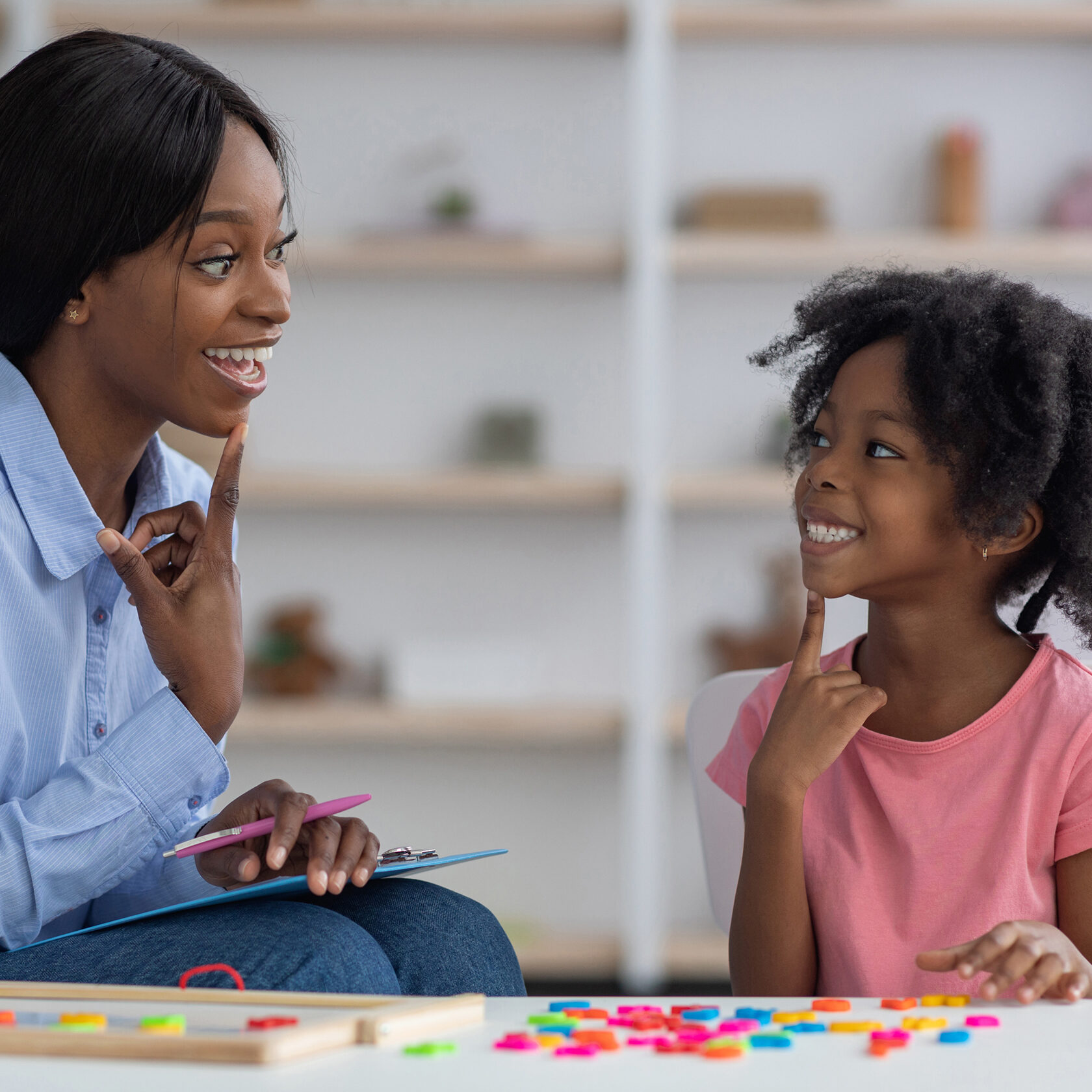 Speech therapist pretty young african american woman working with little black girl with bushy hair at clinic, sitting in front of each other, smiling and touching their chins, side view